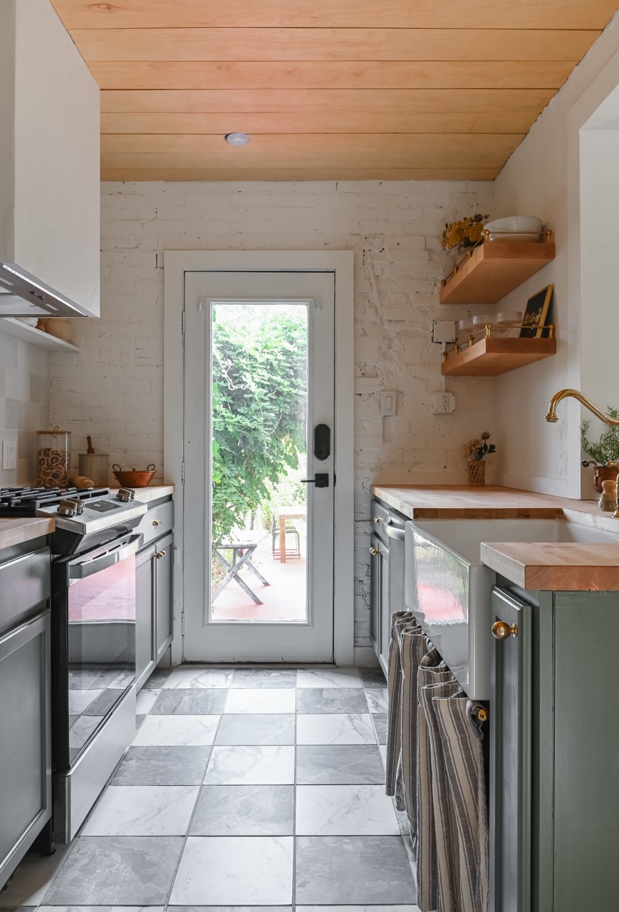 Bright galley kitchen with gray cabinets, wood countertops, checkered gray and white floor tiles, and a glass door opening to a leafy outdoor space.