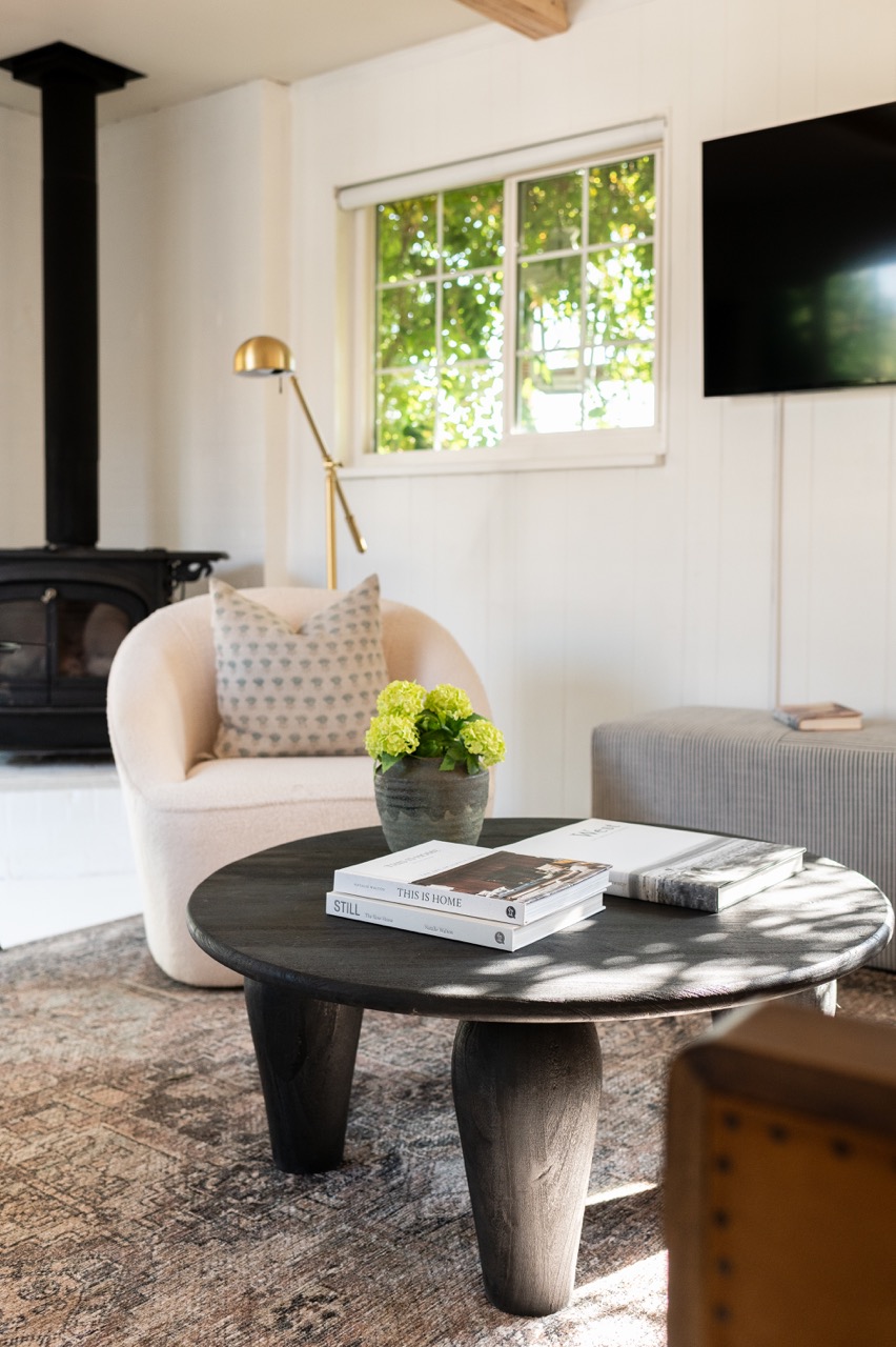 Cozy living room with a round black wooden coffee table holding books and a potted green plant, a beige armchair with a patterned pillow, a floor lamp, and a black wood stove in the background.