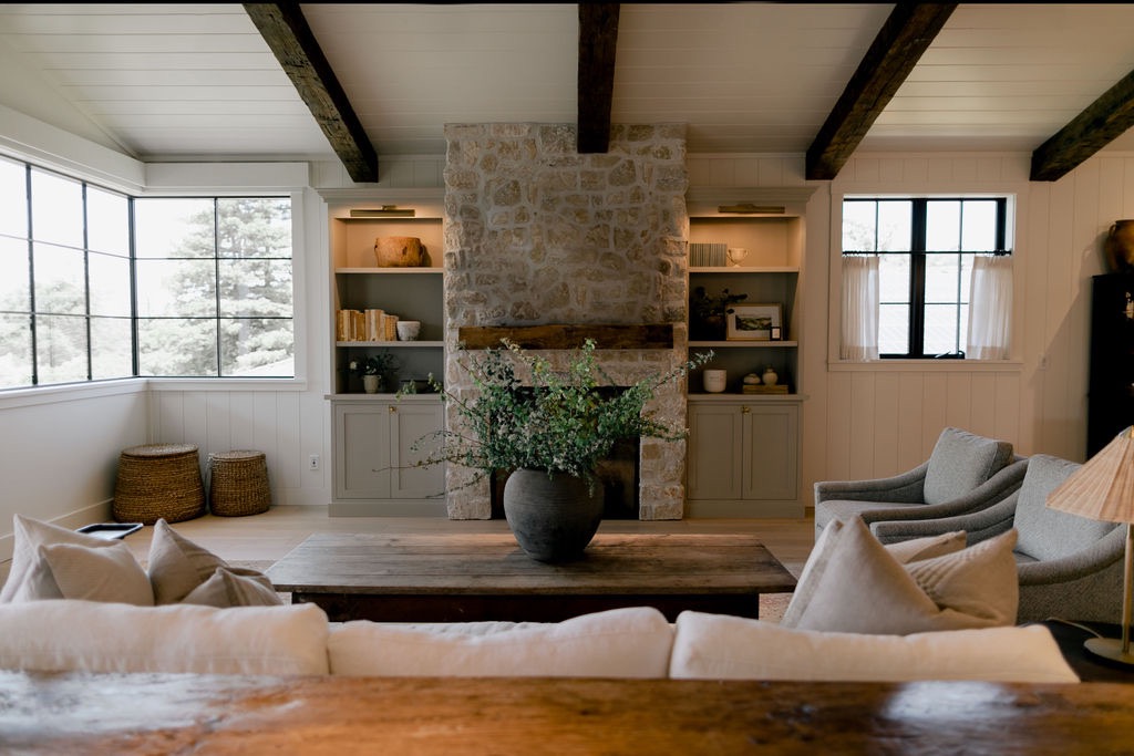 Cozy living room with stone fireplace, wooden coffee table holding a large vase with green branches, gray armchairs, and vaulted ceiling with exposed beams.