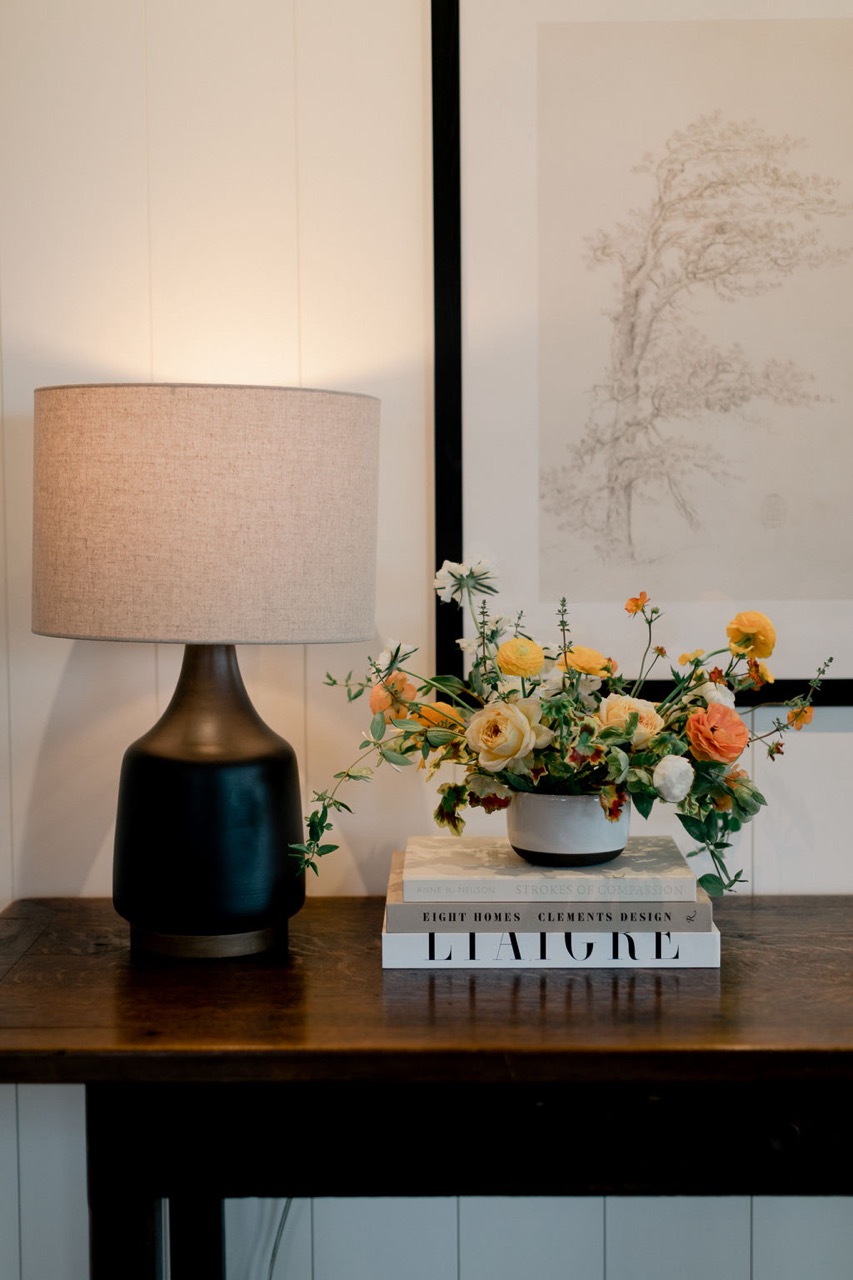 Table lamp with a beige shade next to a vase of yellow and orange flowers on stacked design books atop a wooden console table.