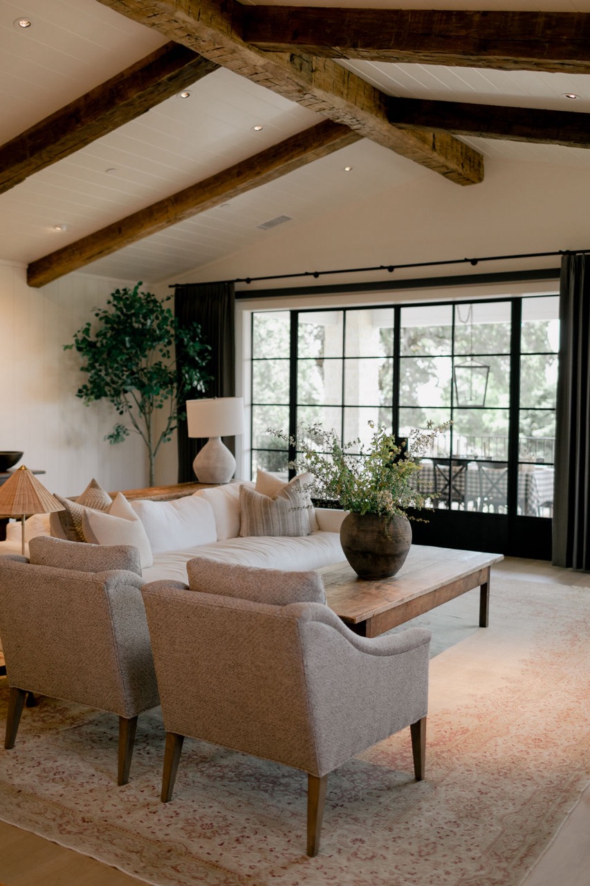 Cozy living room with wooden ceiling beams, a white sofa, two gray armchairs, a wooden coffee table with a vase of greenery, and large black-framed windows.