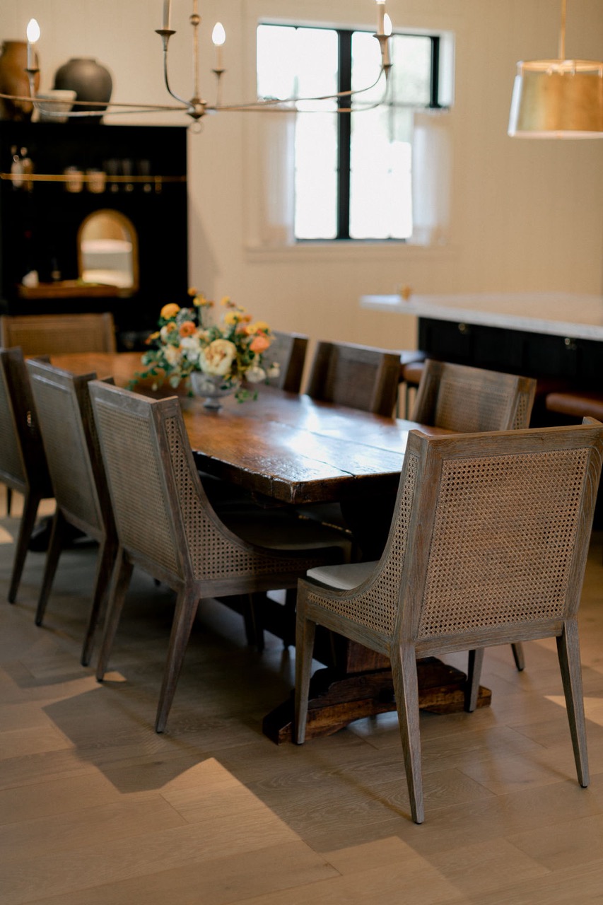 Wooden dining table surrounded by woven back chairs with a floral centerpiece in a softly lit room.