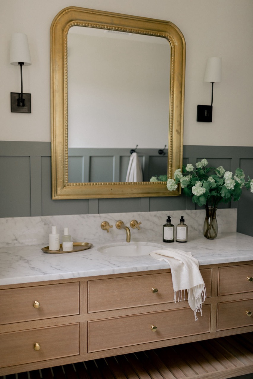Bathroom vanity with marble countertop, gold fixtures, large gold-framed mirror, floral arrangement, and wooden drawers.