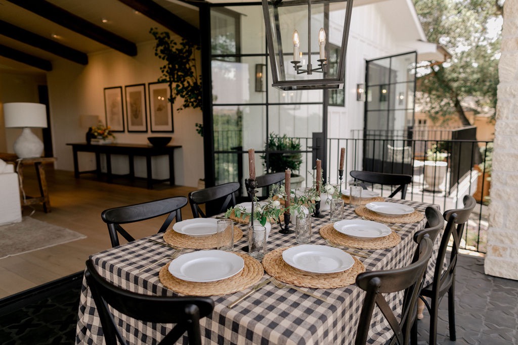Outdoor dining table with black and white checkered tablecloth set with white plates, woven placemats, glassware, candles, and greenery under a hanging lantern light.