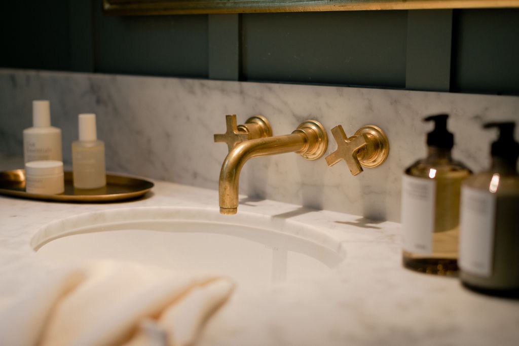 Close-up of a marble bathroom sink with gold-tone wall-mounted faucet and cross handles, with skincare bottles on a tray and soap dispensers nearby.