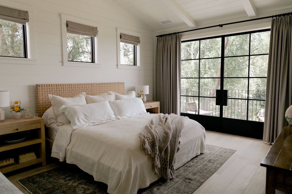 Cozy bedroom with a large bed featuring white linens and a beige throw, wooden nightstands with lamps and flowers, and large glass doors opening to a balcony with trees outside.