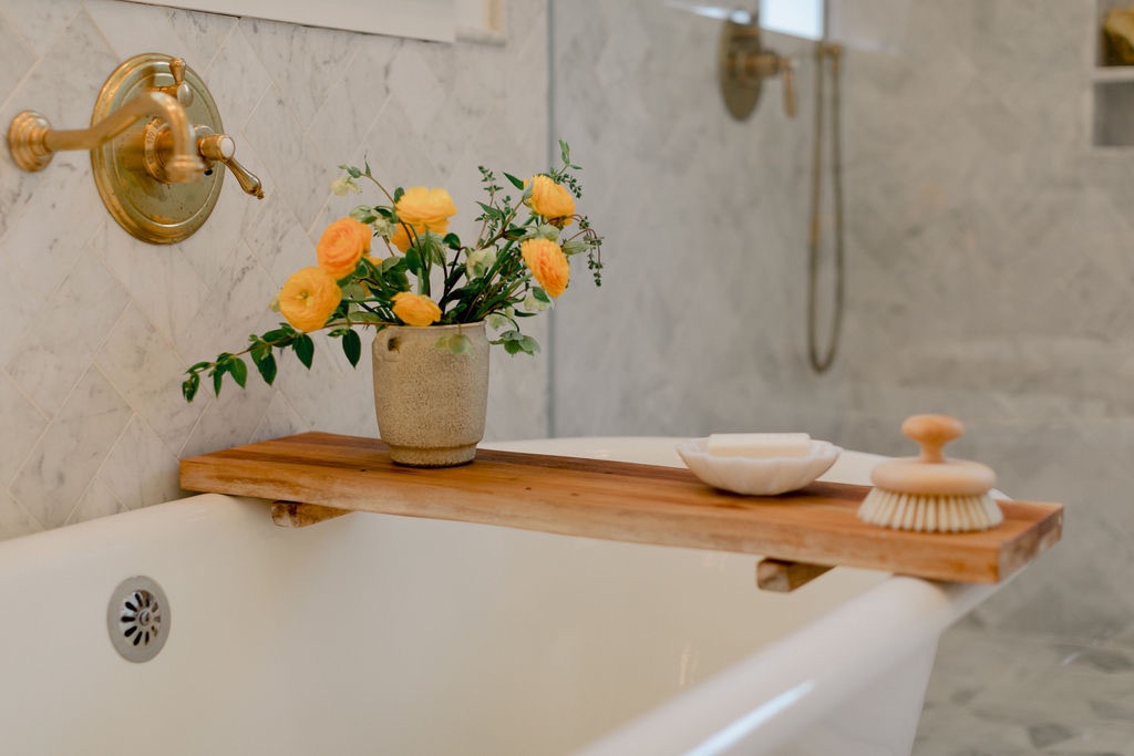 Wooden bath tray across white bathtub holding a vase of yellow flowers, soap dish, and wooden brush, with brass faucet and marble tiles in background.