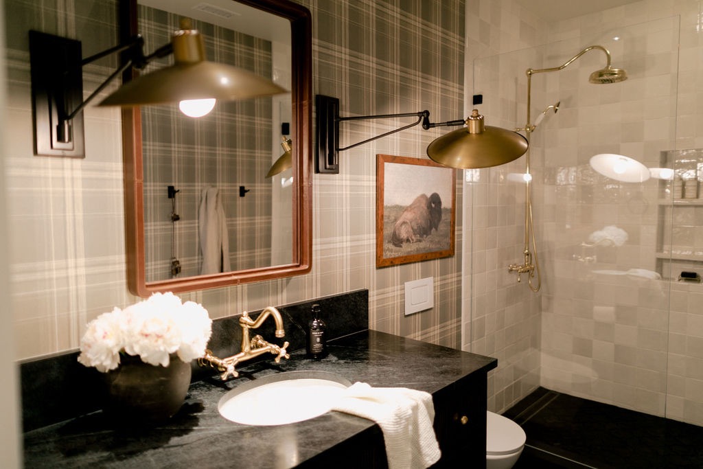 Bathroom with black countertop, brass faucet, white towel, wall-mounted brass light, framed bison picture, and brass shower fixtures.