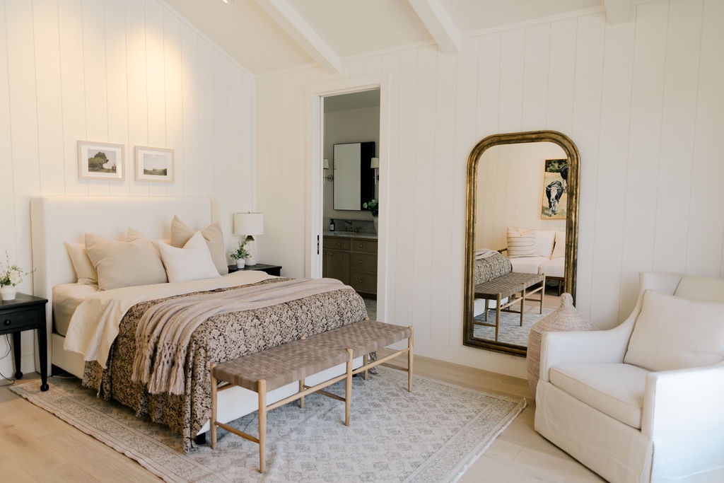 Bright bedroom with a white upholstered bed, beige and white pillows, patterned blanket, bench at the foot, white armchair, and large arched mirror reflecting part of the room.