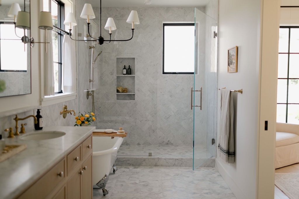 Light and airy bathroom with marble walls and floor, featuring a clawfoot bathtub, glass-enclosed shower, wooden vanity, and brass fixtures.