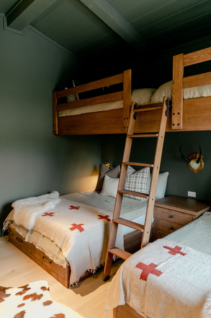 Cozy bedroom with a wooden bunk bed, ladder, twin beds with gray bedding, and blankets featuring red crosses.