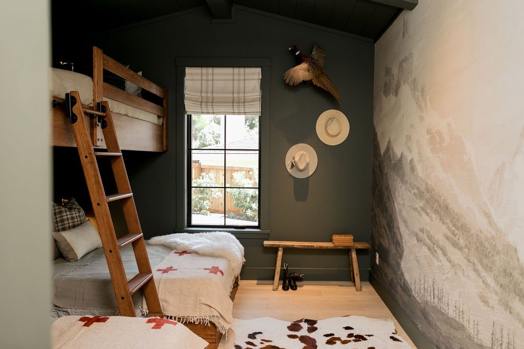 Cozy bedroom with wooden bunk beds, a window with plaid curtain, two hats on dark green wall, mounted pheasant, rustic bench, and mountain mural.