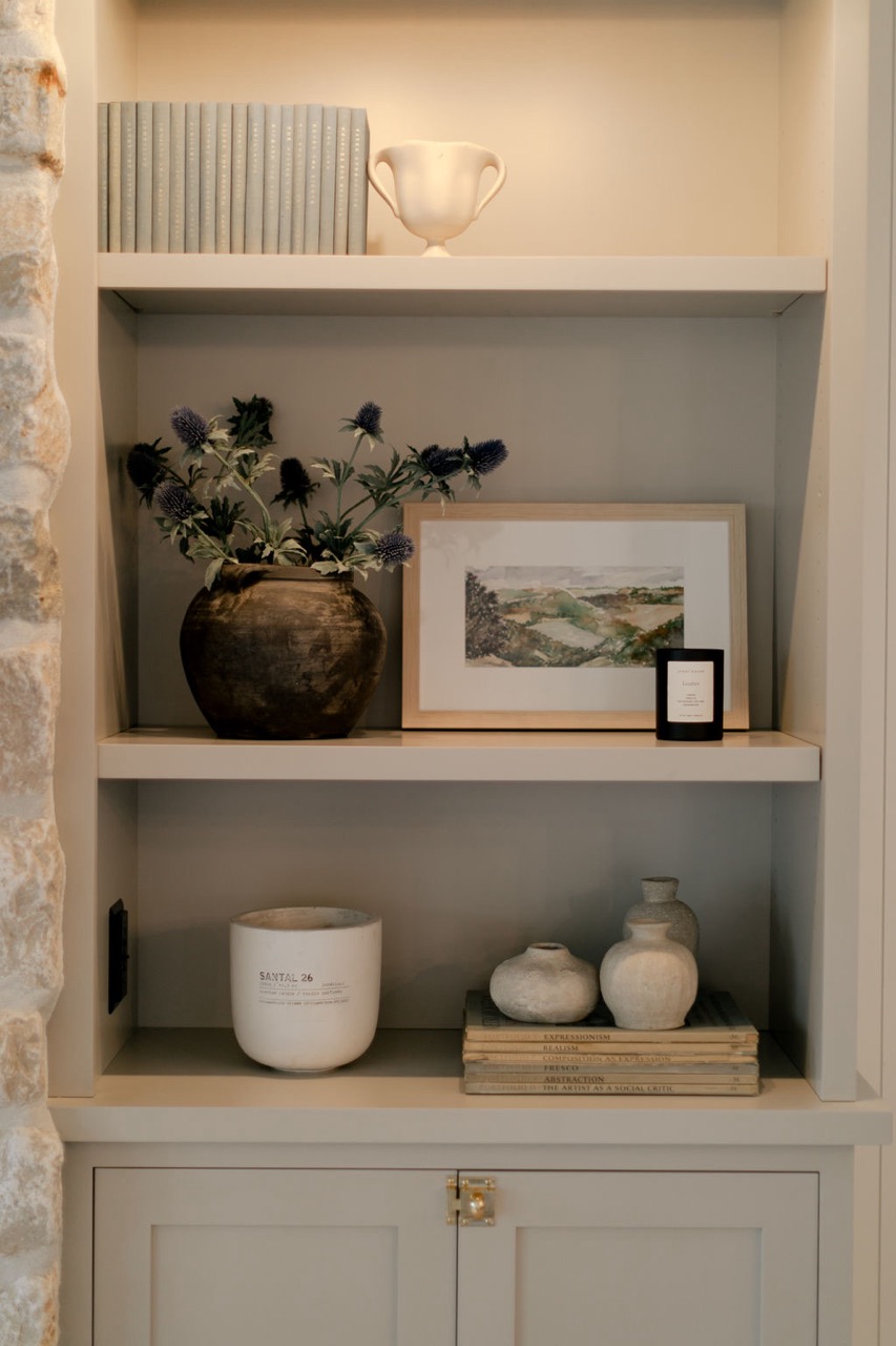 White built-in shelves displaying decorative items including a row of grey books, a white trophy-shaped vase, a dark brown vase with purple flowers, a framed landscape painting, a black candle, a white candle labeled Santal 26, and three small beige pottery vases on top of stacked books.