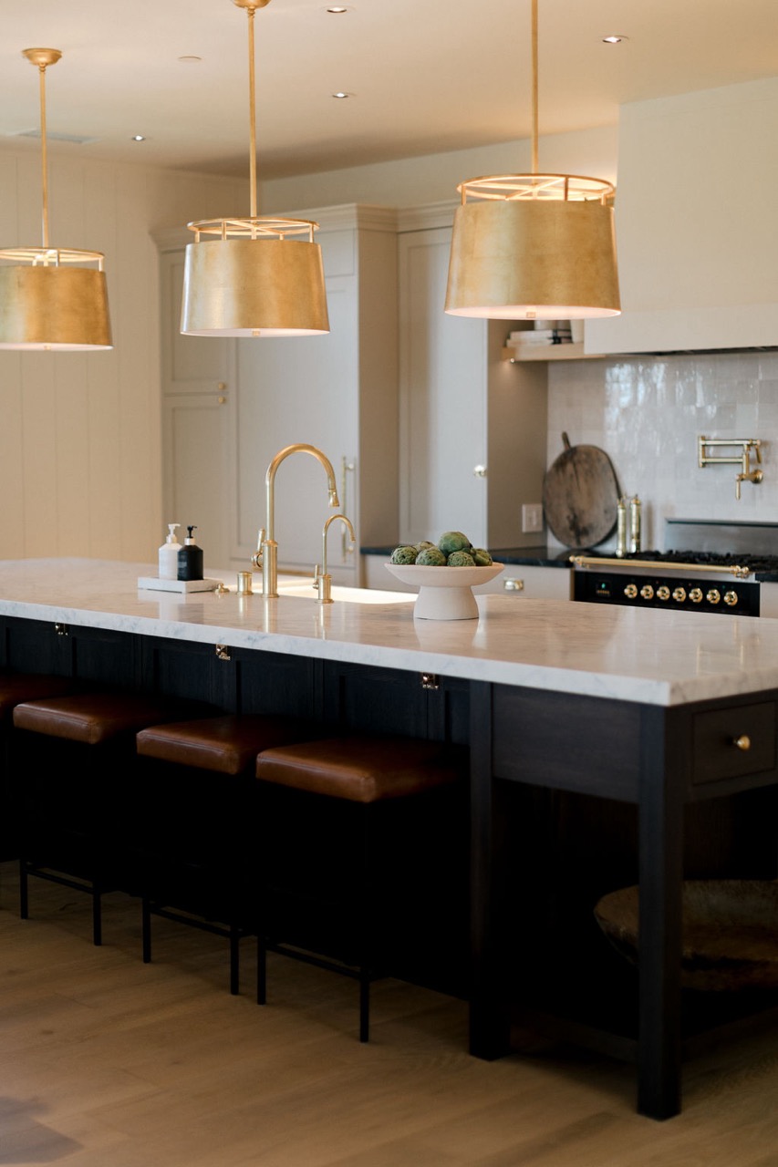Modern kitchen island with a white marble countertop, gold pendant lights, gold faucets, brown leather stools, and a bowl of artichokes.
