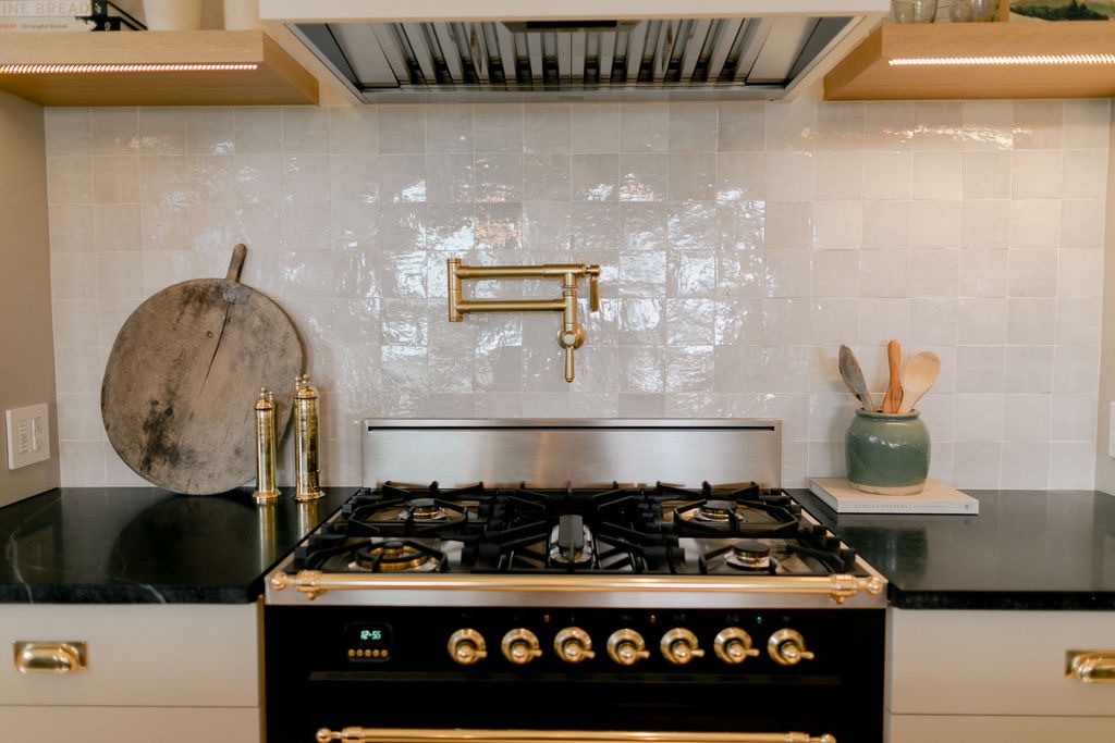 Modern kitchen stove with brass knobs, a mounted brass pot filler faucet, and black countertops with cooking utensils and cutting board.