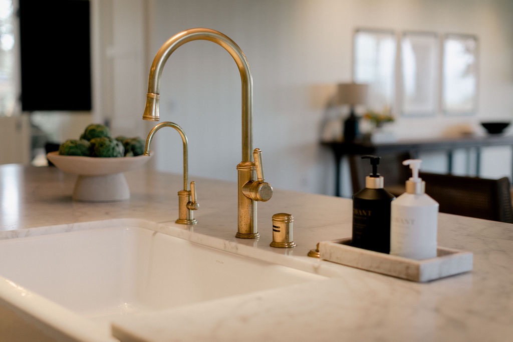 Close-up of a kitchen sink with gold-toned faucet and soap dispensers on a marble countertop.