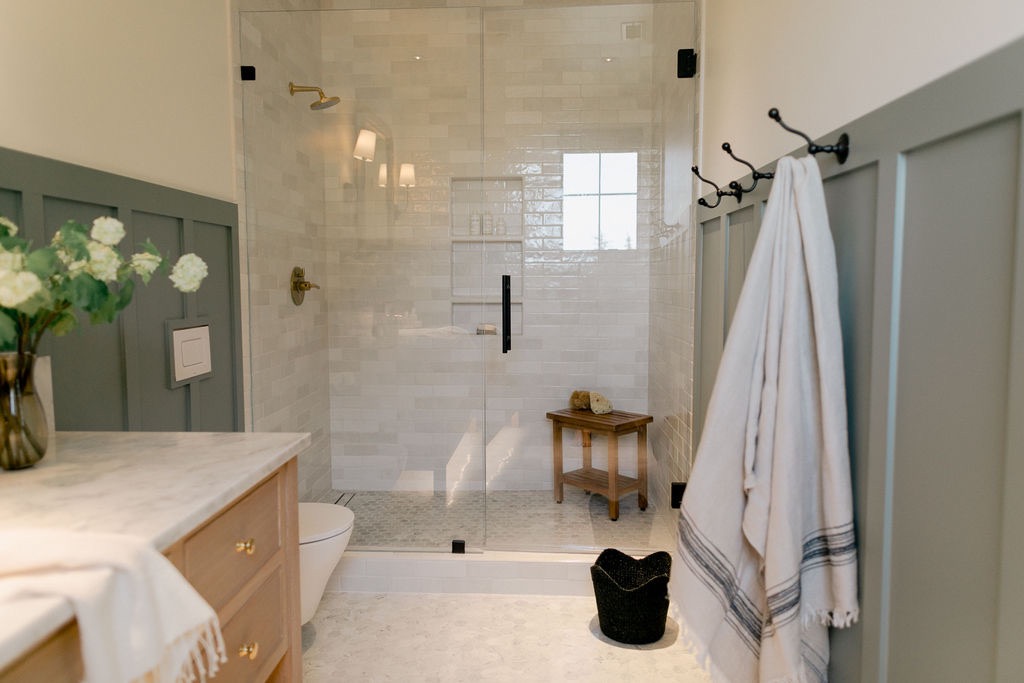 Modern bathroom with glass-enclosed shower, light wood cabinetry, marble countertop, white and gray towels hanging on hooks, and a small wooden stool inside the shower.