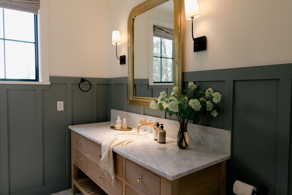 Bathroom vanity with marble countertop, gold faucet, a large gold-framed mirror, wall sconces, and a vase of white flowers.