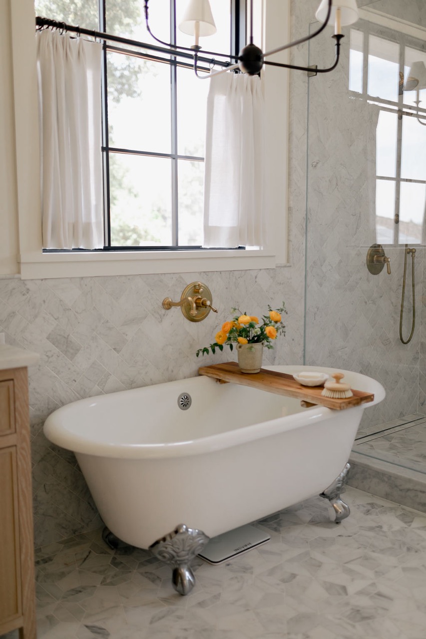 White clawfoot bathtub with wooden tray holding a vase of yellow flowers and bath accessories in a marble-tiled bathroom with a window and curtains.