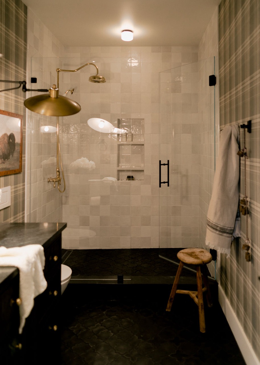 Bathroom with a glass-enclosed shower featuring white tiled walls, brass showerhead, plaid wallpaper, wooden stool, and dark floor tiles.