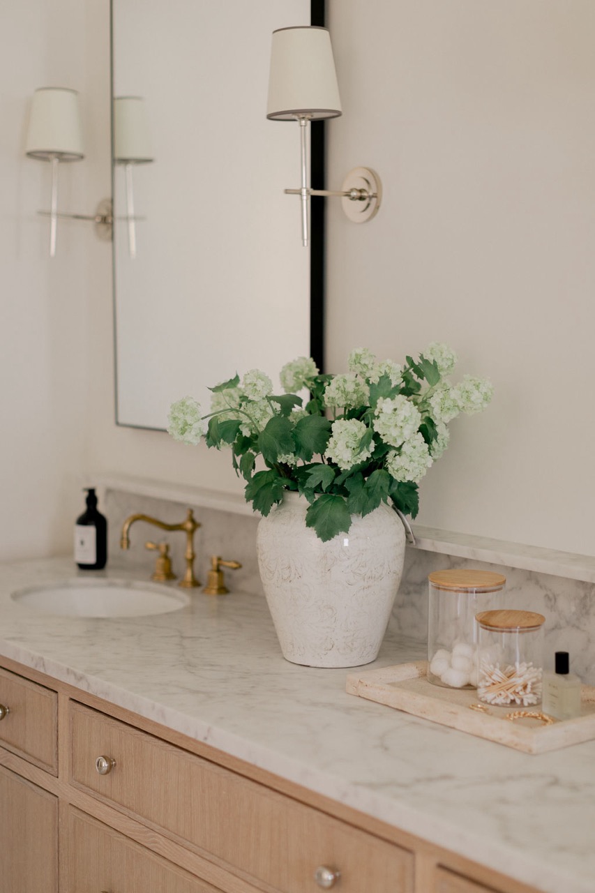 Bathroom vanity with marble countertop, brass faucet, white vase with green and white flowers, glass containers, and wall-mounted lamp next to a mirror.