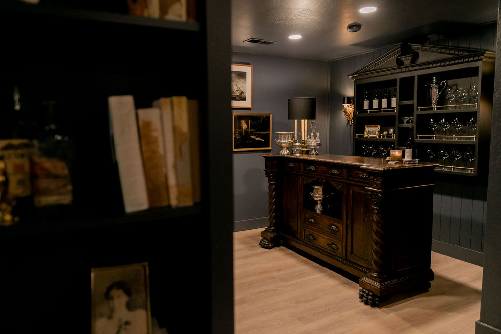 Dimly lit room with an ornate wooden bar cabinet, glassware, and framed pictures on dark walls.