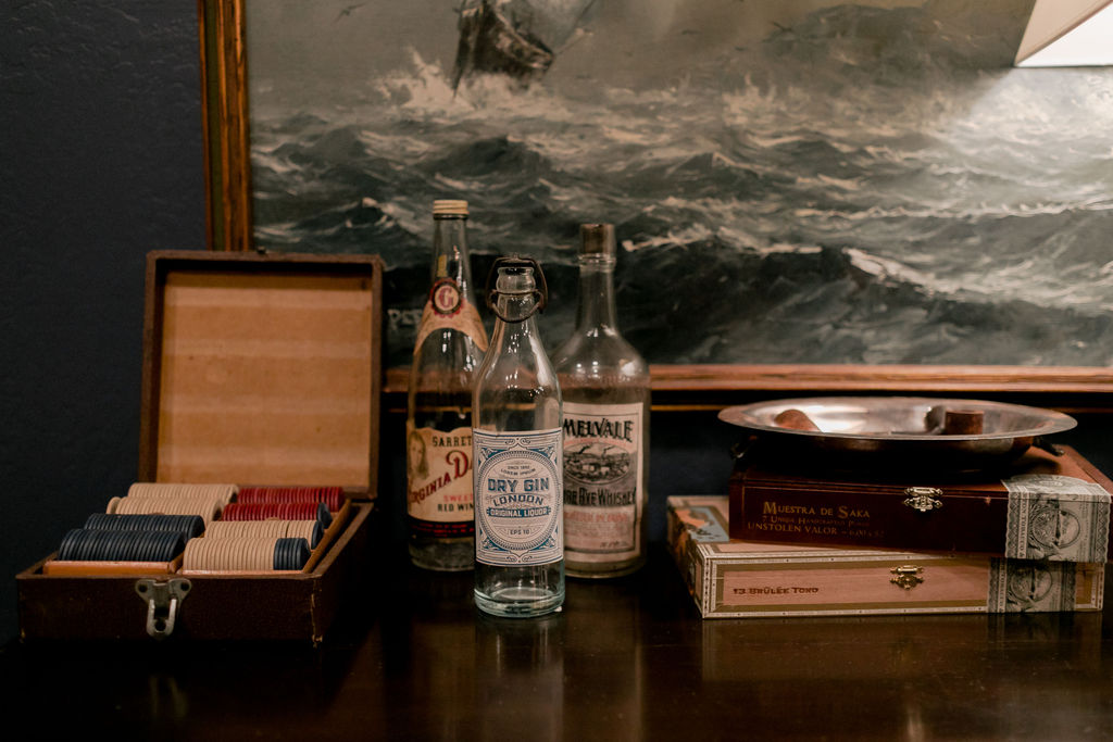 Dark wooden table with vintage liquor bottles, an open box of poker chips, and stacked cigar boxes under a framed seascape painting.