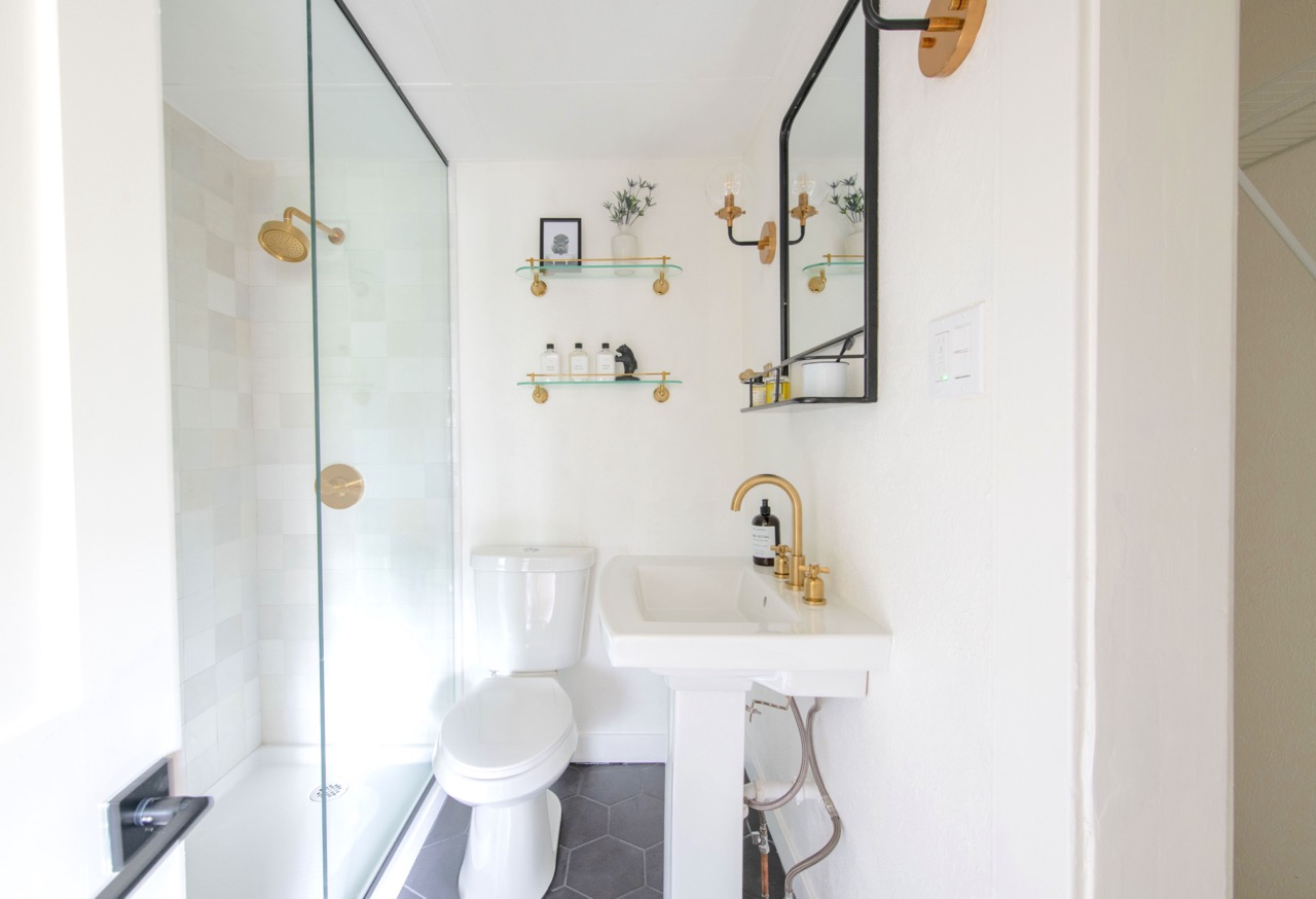 Bright white bathroom with glass-enclosed shower, white toilet, pedestal sink with gold faucet, black-framed mirror, and gold-accented shelves holding toiletries and decor.