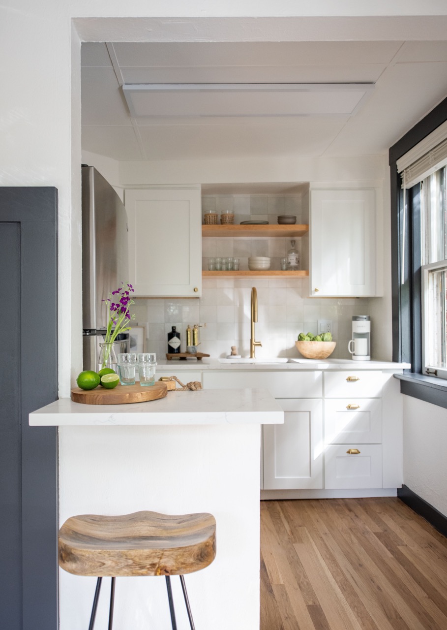 Bright kitchen with white cabinets, wooden floor, a wooden stool, and a white countertop displaying a wooden tray with limes, glasses, and purple flowers in a vase.