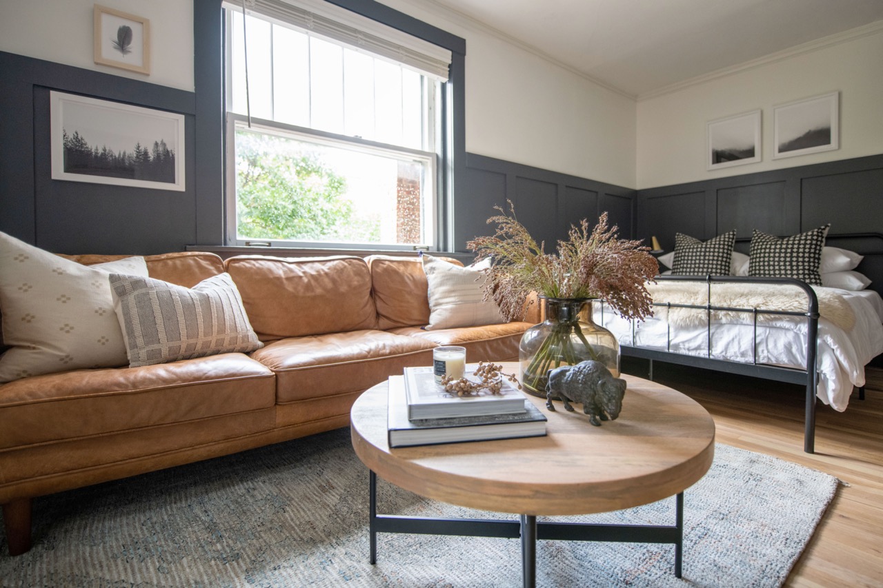 Cozy living room with a caramel leather couch, round wooden coffee table with books and a vase of dried flowers, and a black metal bed with white bedding and patterned pillows.
