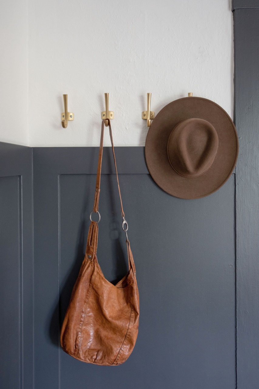 Three gold coat hooks mounted on a white and dark gray wall, holding a brown leather bag and a brown felt hat.
