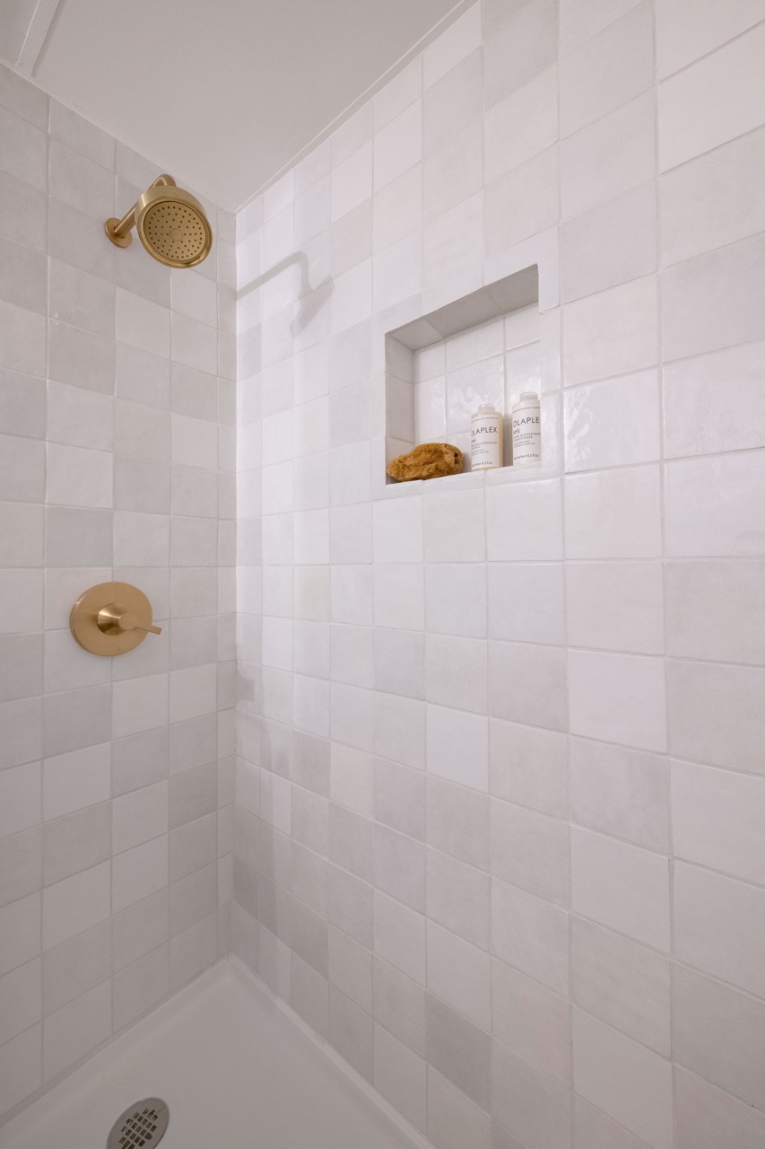 Shower with white square tiled walls, gold showerhead and handle, and a recessed shelf holding two bottles and a brown sponge.