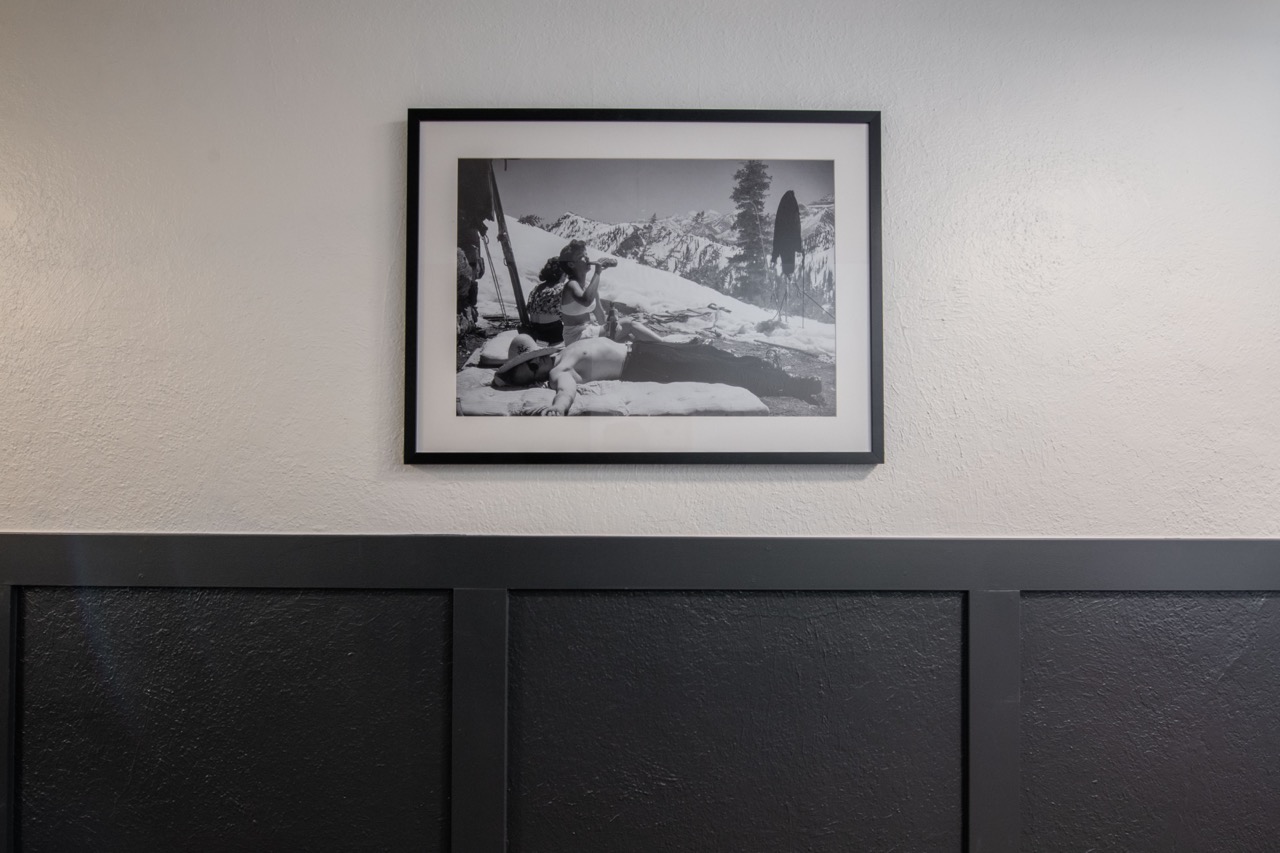 Shelf with bottles of liquor, glassware, a vase with white flowers, lemons on a cutting board, and a framed forest artwork, with stacks of books below.