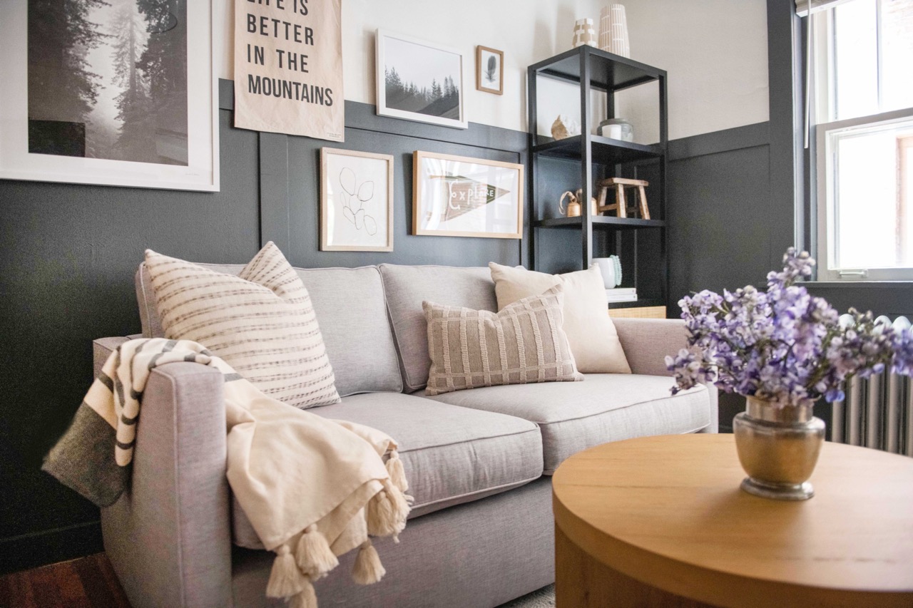 Cozy living room with a light gray sofa adorned with neutral throw pillows and blankets, a wooden round coffee table with a vase of purple flowers, and wall art including a banner that reads 'Life is better in the mountains.'