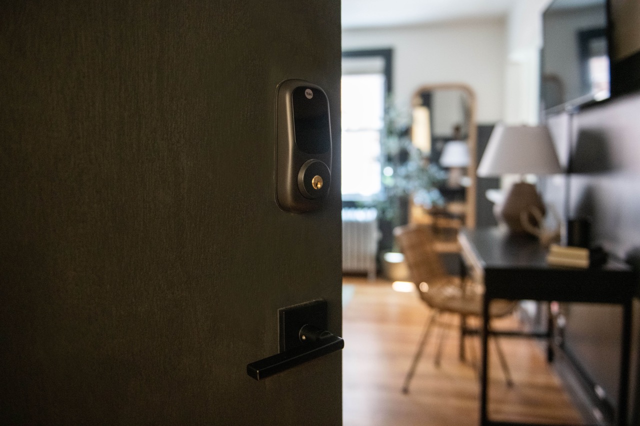 Partially open dark wooden door with a modern black electronic lock, revealing a blurred view of a cozy, well-lit room with a desk, chair, and lamp.