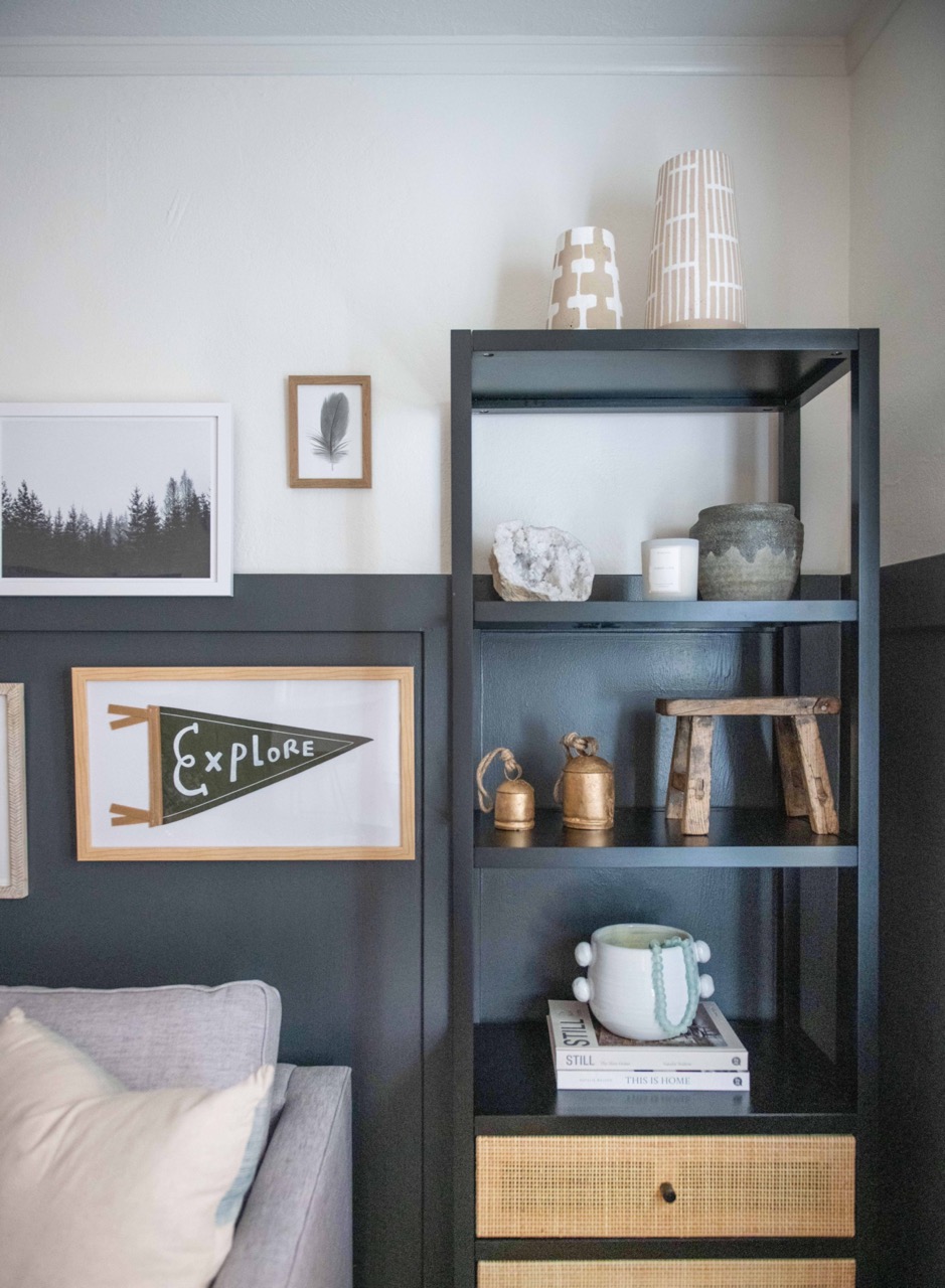 Black shelving unit with decorative vases, wooden stool, brass bells, a white candle, a stone, and a white pot with a beaded necklace, next to framed wall art and a gray couch with a white pillow.