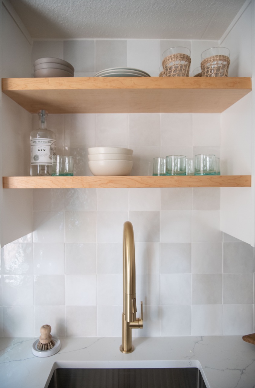 Modern kitchen sink with a tall brushed gold faucet, light wood racks above holding bowls, glasses, and a gin bottle against a tiled backsplash.