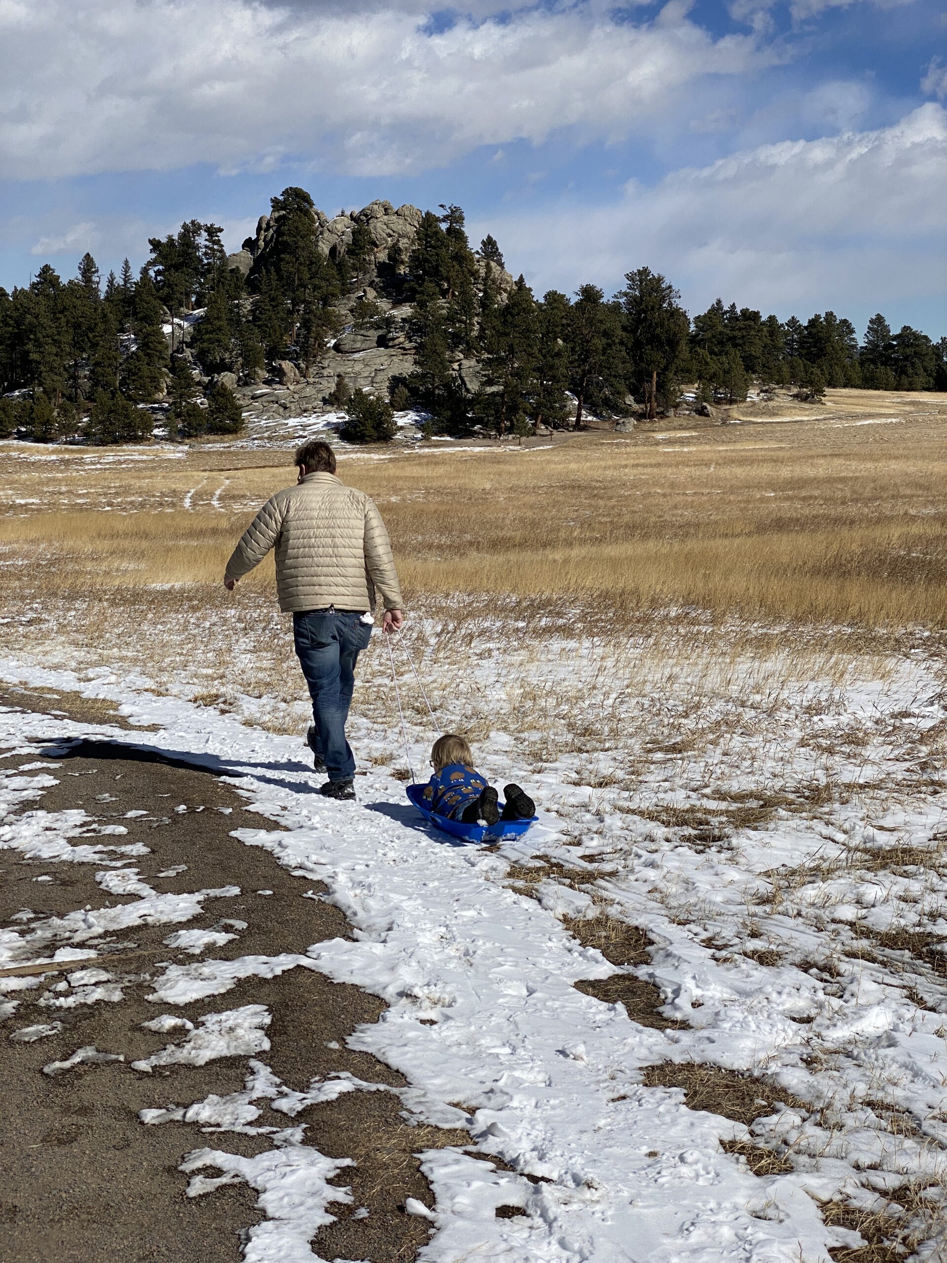 Person pulling a small child lying on a blue sled along a snowy path in an open field with pine trees and rocky hills in the background.