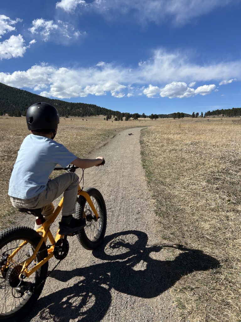 Child wearing a helmet riding a yellow bike on a dirt path through a dry grassy field under a blue sky with scattered clouds.