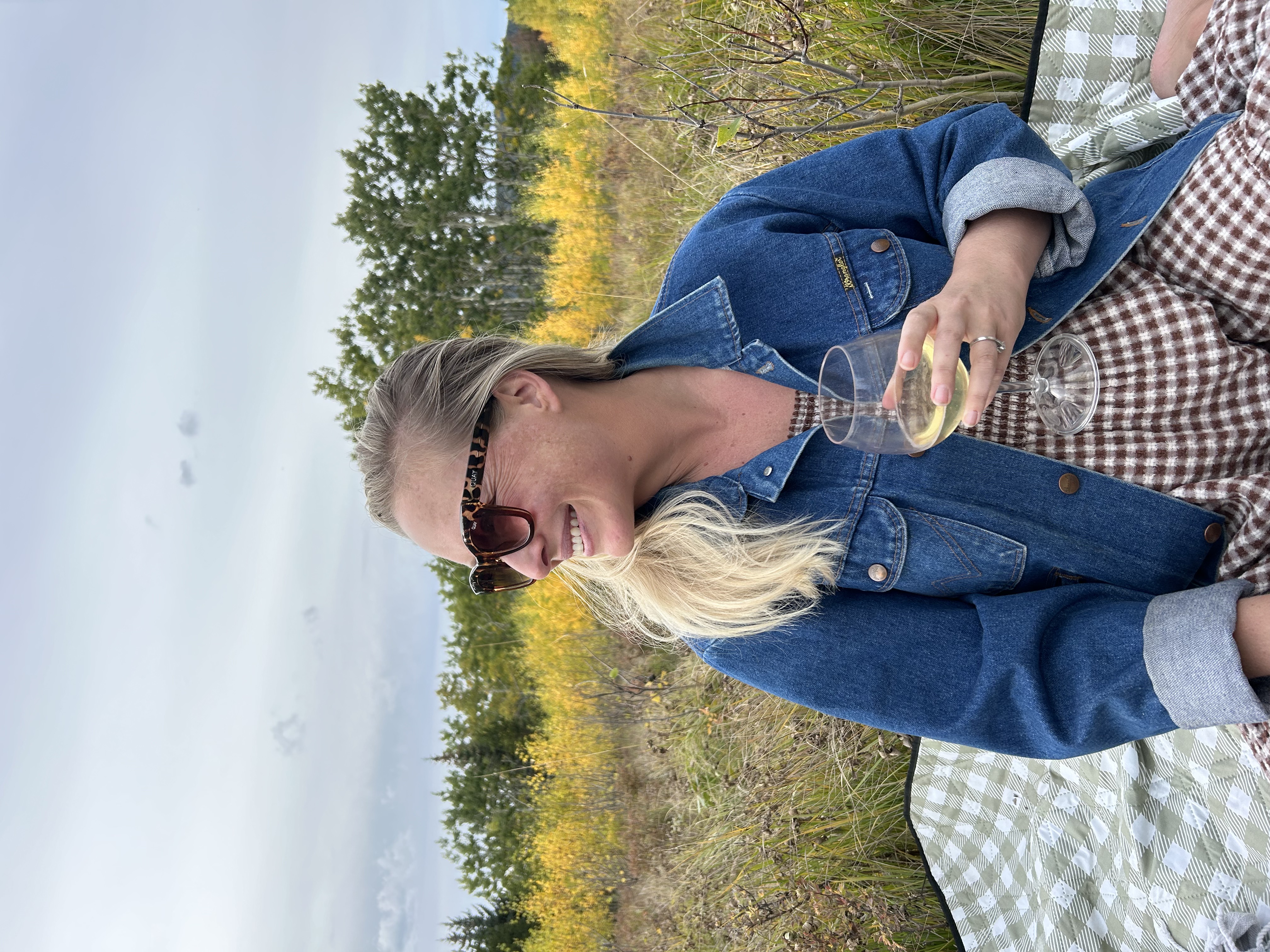 Smiling woman in a denim jacket and sunglasses holding a glass of white wine sitting on a checkered blanket in a grassy field with trees in the background.