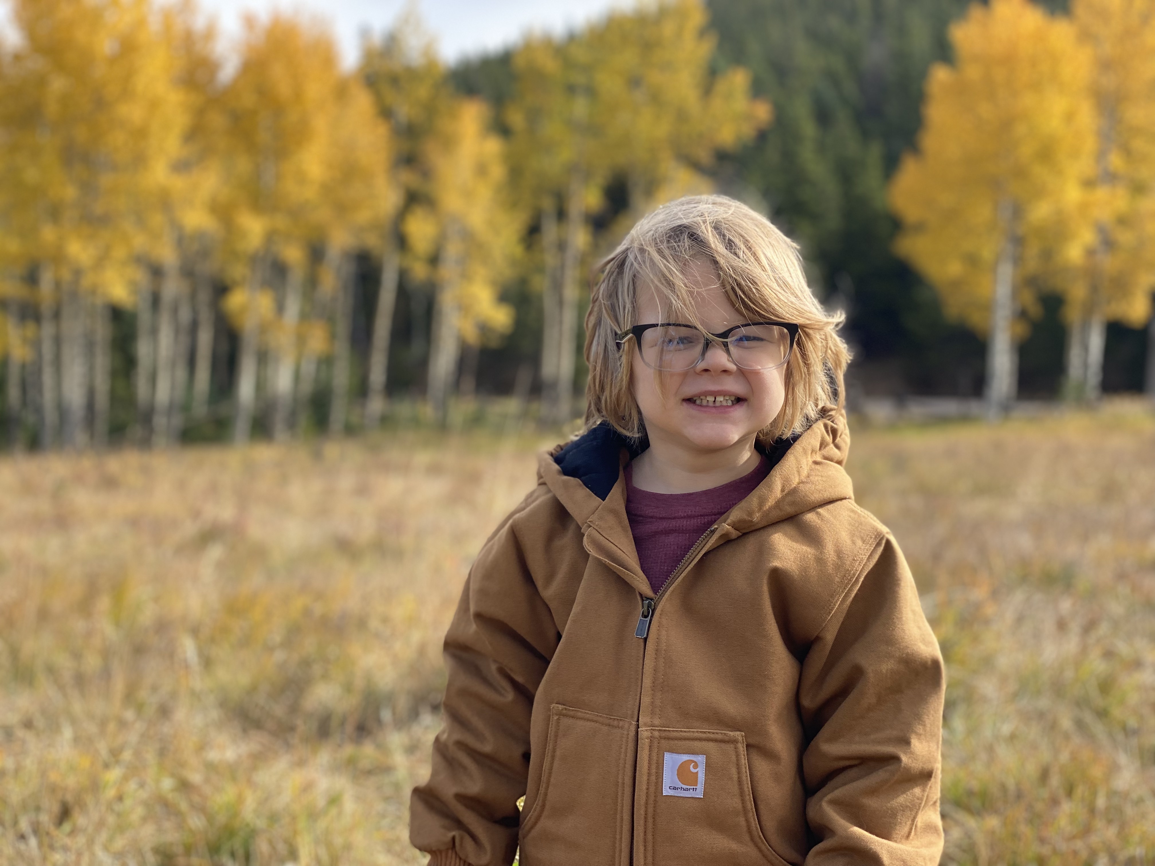 Smiling child wearing glasses and a brown jacket standing in a field with yellow autumn trees in the background.