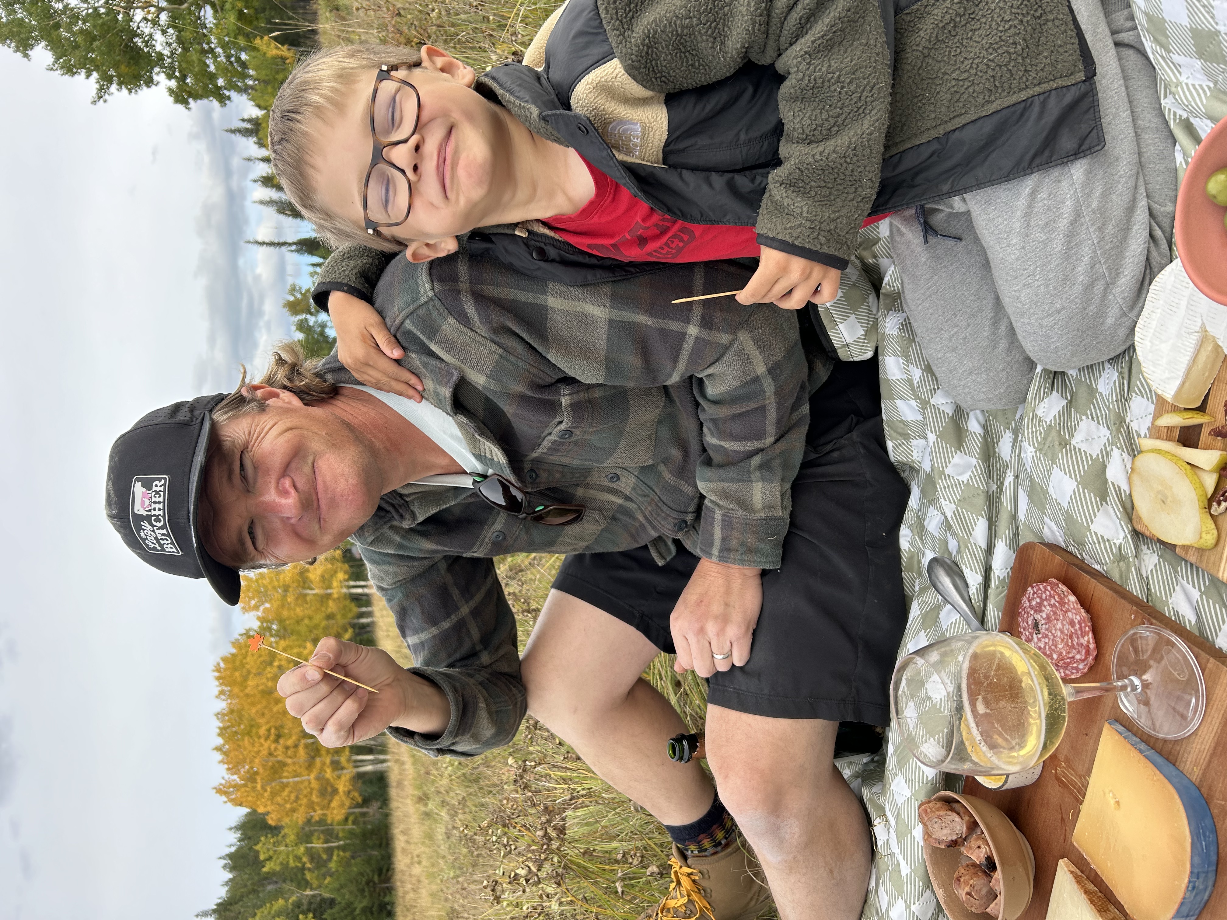 Man wearing a black cap and plaid shirt sitting on a blanket outdoors with a young boy in glasses and a fleece jacket, enjoying a picnic with cheese, sausages, salami, sliced pears, green olives, and a glass of white wine.