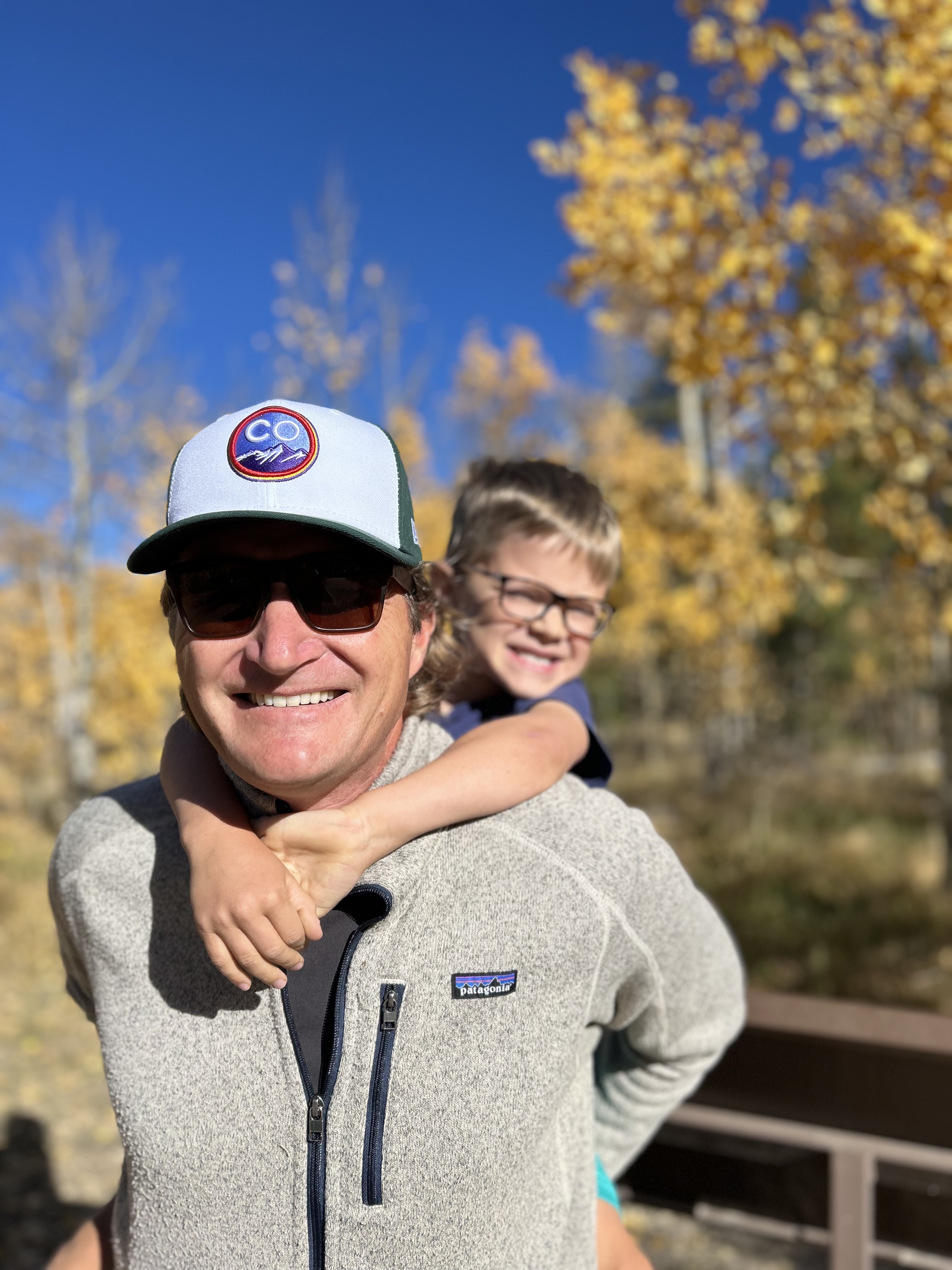 Smiling man wearing sunglasses and a hat with a child giving him a piggyback ride outdoors with autumn yellow trees in the background.