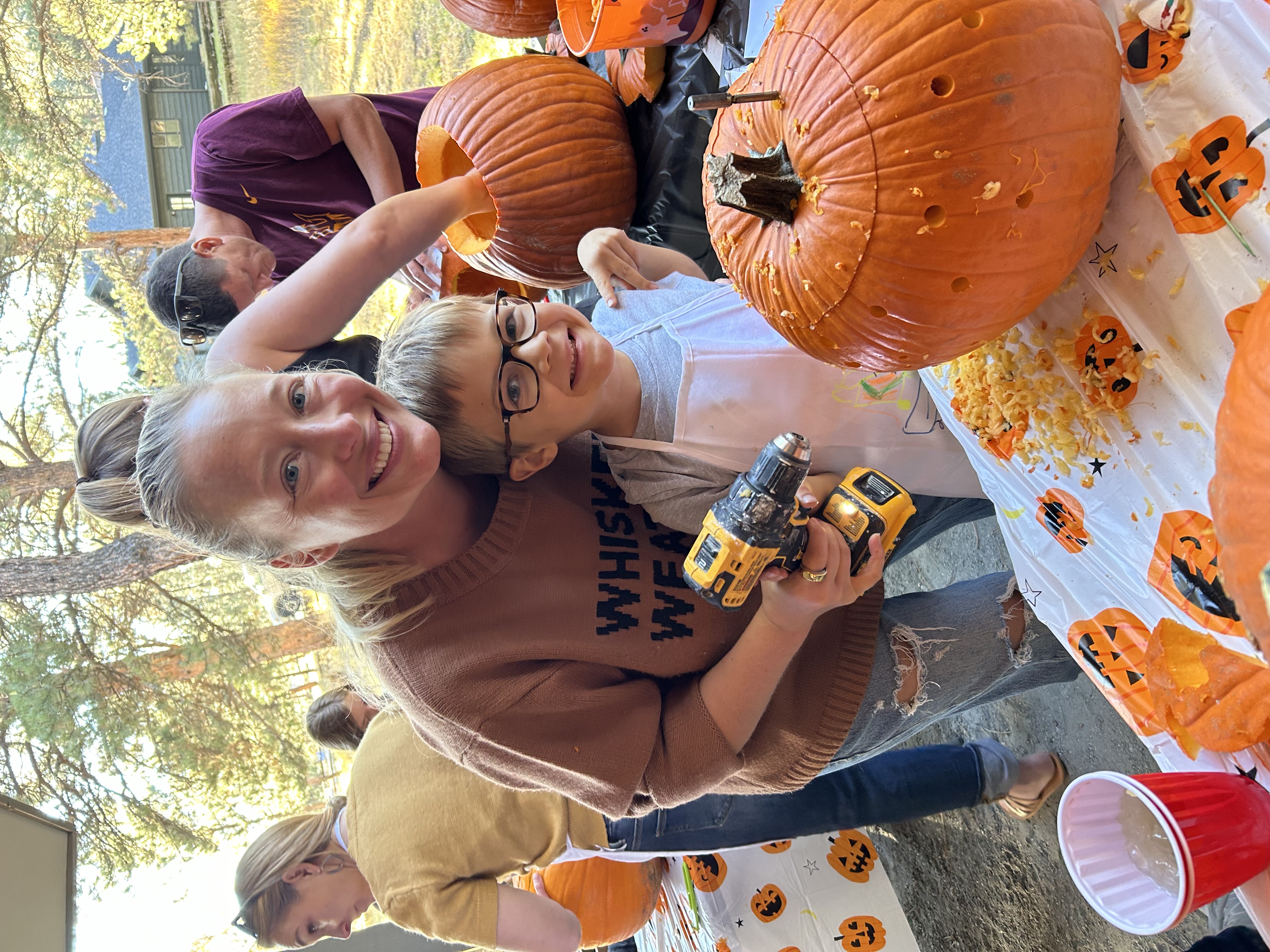 Smiling woman and young boy holding a drill while carving a pumpkin at an outdoor pumpkin carving event.