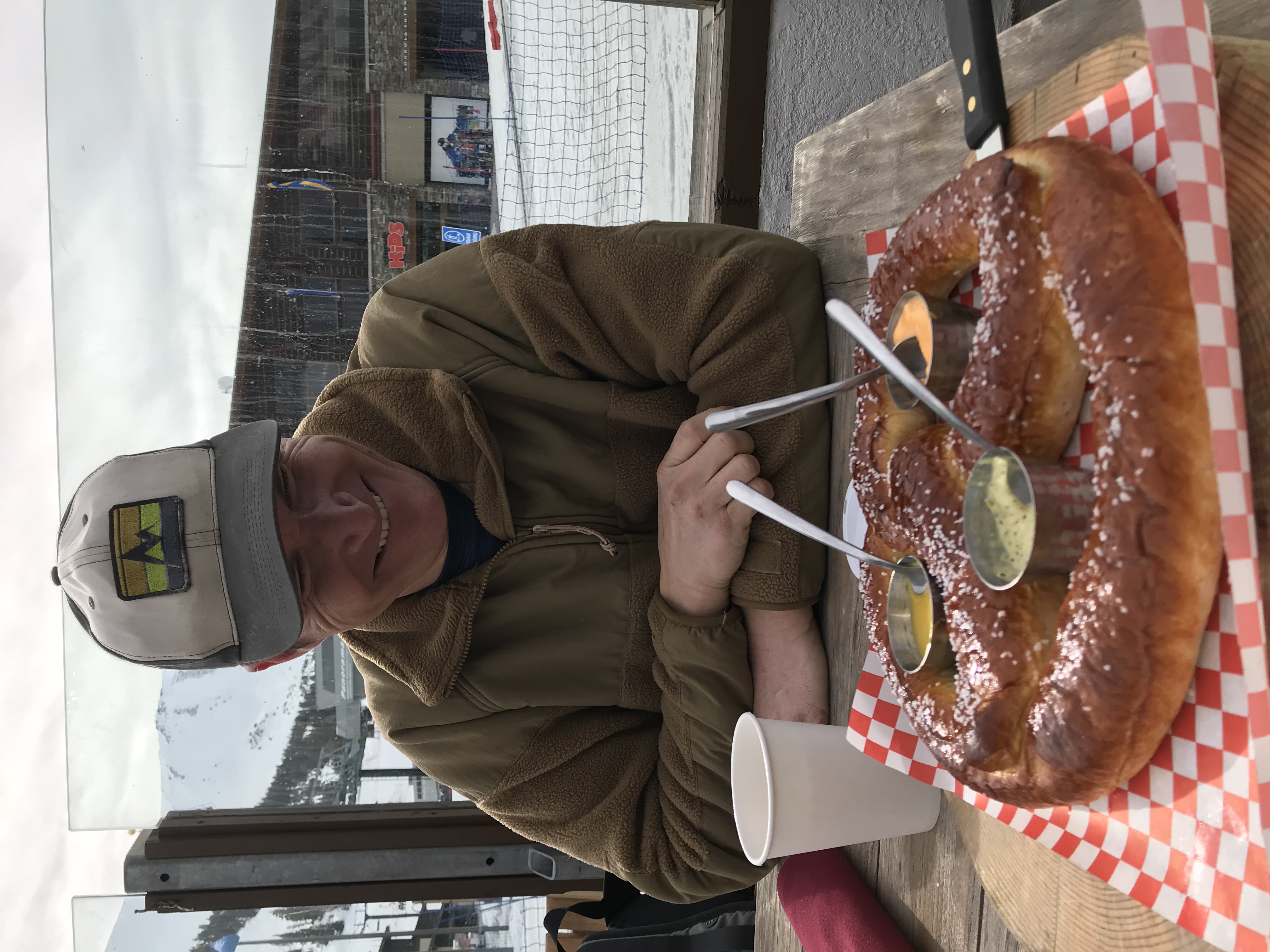 Smiling man in a brown jacket and cap sitting at a wooden table with a large soft pretzel and three dipping sauces in front of him, with a snowy mountain scene visible through the window behind.