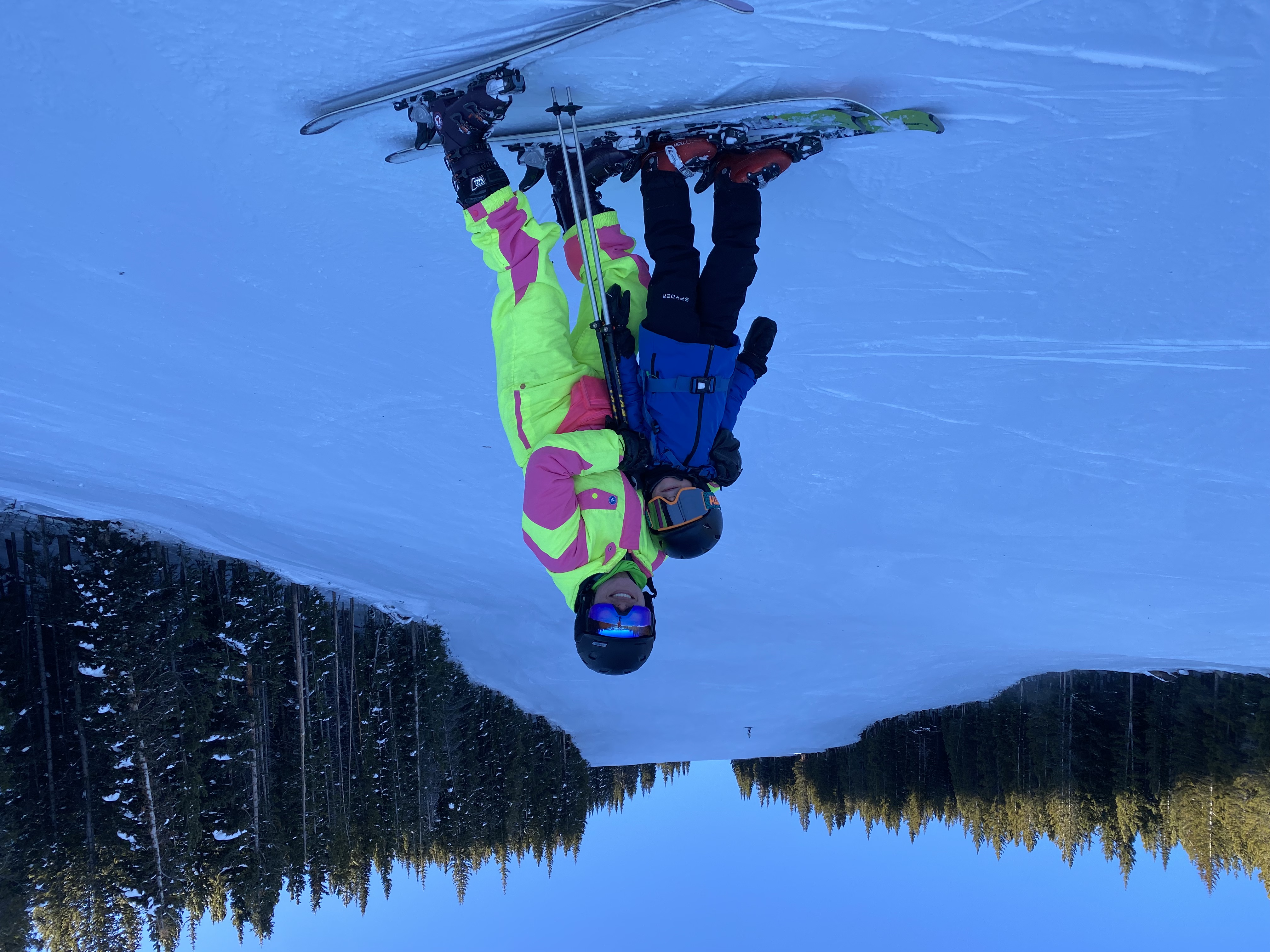 Adult and child posing together on skis on a snowy slope surrounded by pine trees under a clear blue sky.