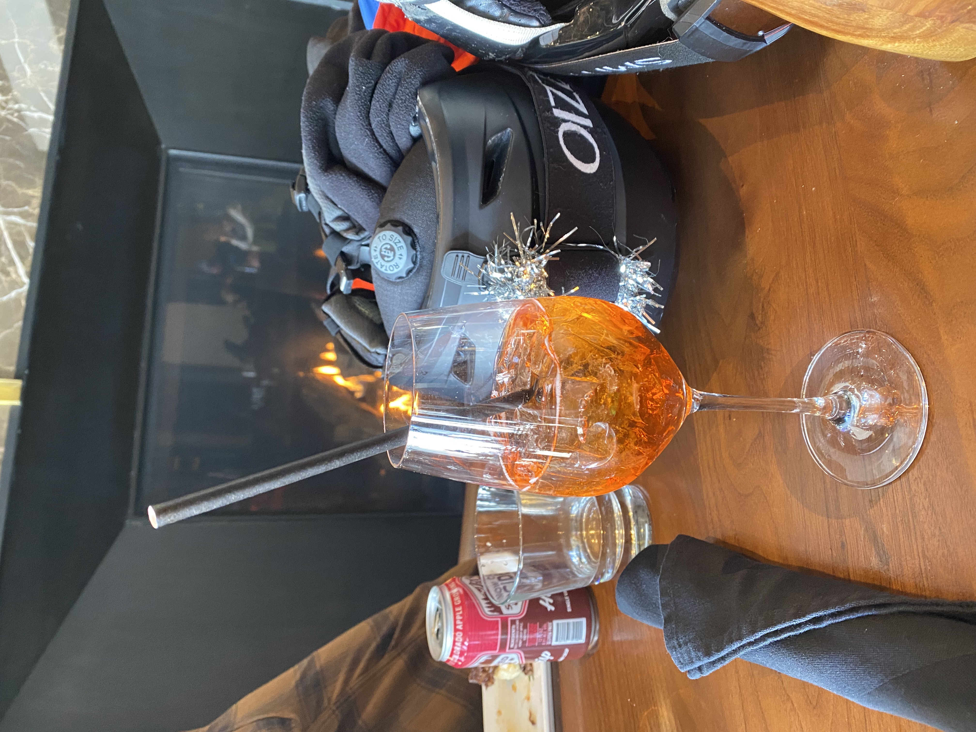 Glass of orange cocktail with ice and black straw on wooden table near black napkin, cycling helmets, and a fireplace in background.