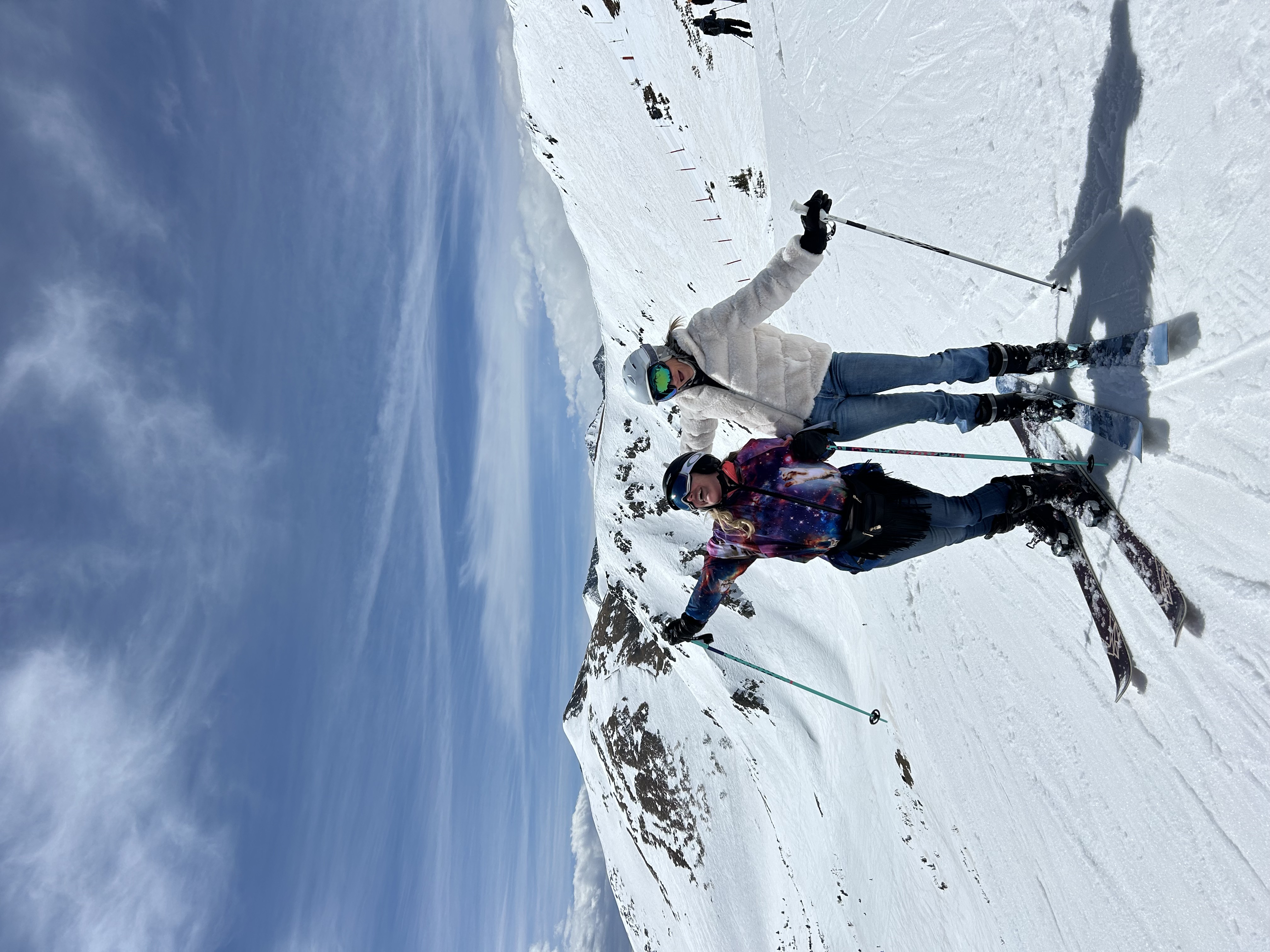 Two women in ski gear posing happily on a snowy mountain slope with blue sky and snow-covered peaks in the background.