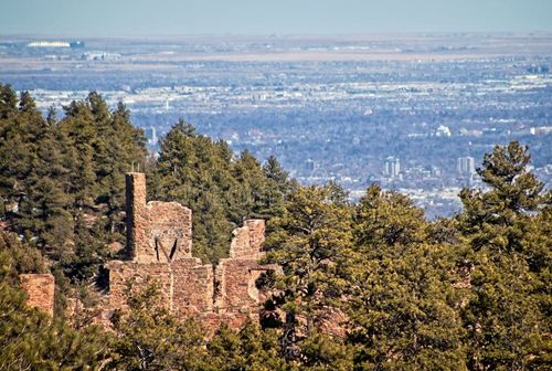 Ruins of a stone building partially hidden among evergreen trees overlooking a distant cityscape under a clear sky on Mt. Falcon in Morrison, CO.