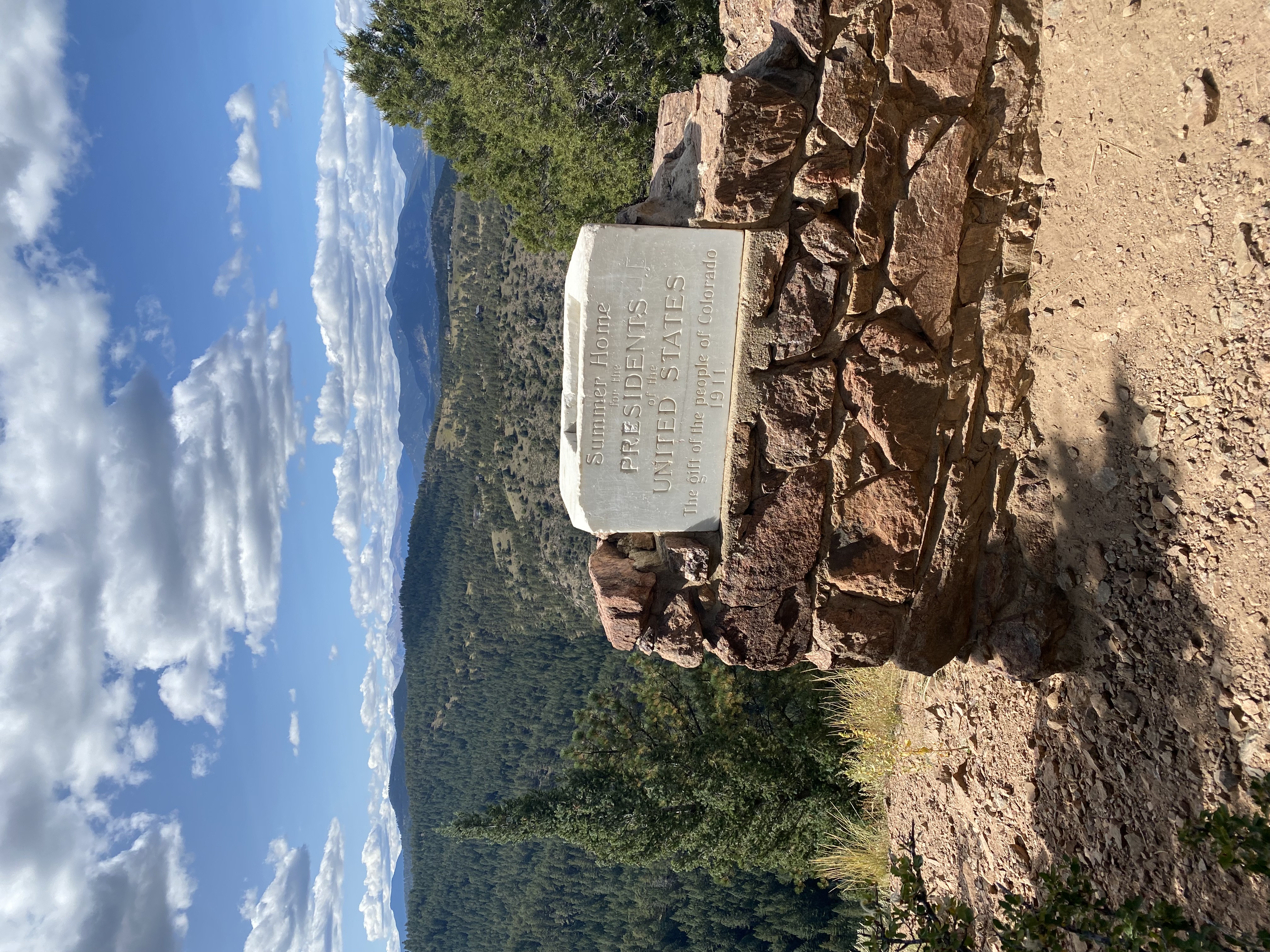 Stone monument with an engraved plaque reading 'Summer Home for the Presidents of the United States, The gift of the people of Colorado 1911' with forested mountains and blue sky with clouds in the background on Mt. Falcon.